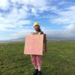 Figure standing field with blue sky wearing a pink sandwich board and yellow hat
