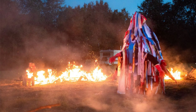 Image of hooded and costumer figure surrounded by small fires at dusk, with trees in the background