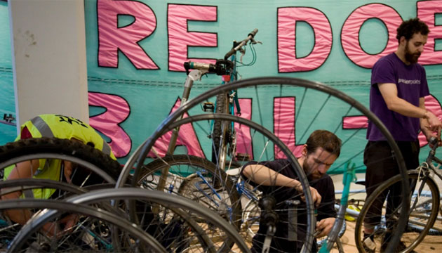 Two men working on bike maintenance, in front of a pink on green banner saying "Nature Doesn't Do Bailouts"