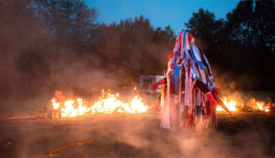 hooded and costumed figure surrounded by small fires, at dusk, with trees in background
