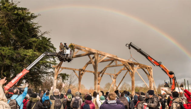 The framework of a barn being raised by a large group of people. Trees and a rainbow can be seen in the background