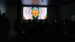 darkened room with large film screen on end wall showing colourful head image