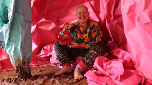 colour photo, interior. Kitt, a white, shaven headed person with a wide crinkly nosed smile, sits amongst vast sheets of pink and mint green paper crumple to form a cave -like structure around them. on the floor is wet, raw terracotta clay in rolling folds