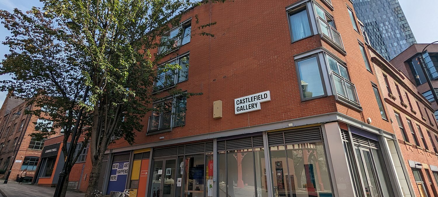 Exterior view of Castlefield Gallery, a modern red-brick building with large windows and the gallery’s name displayed on a sign above its entrance. Bicycles are parked outside and trees line the pavement.