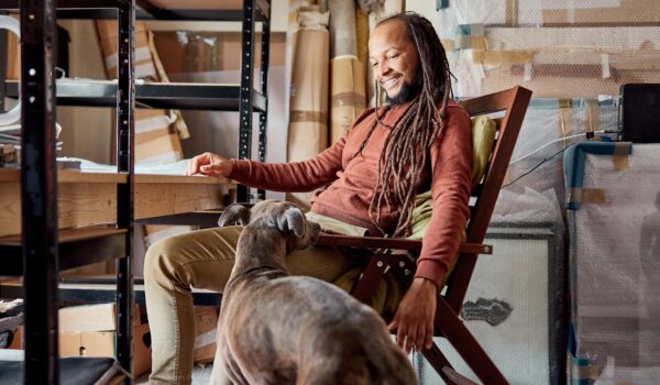 Artist Amartey Golding sat in a wooden armchair, surrounded by a studio-type set up- shelves full of working materials. He has a happy expression as the dog sniffs around his feet at the table.