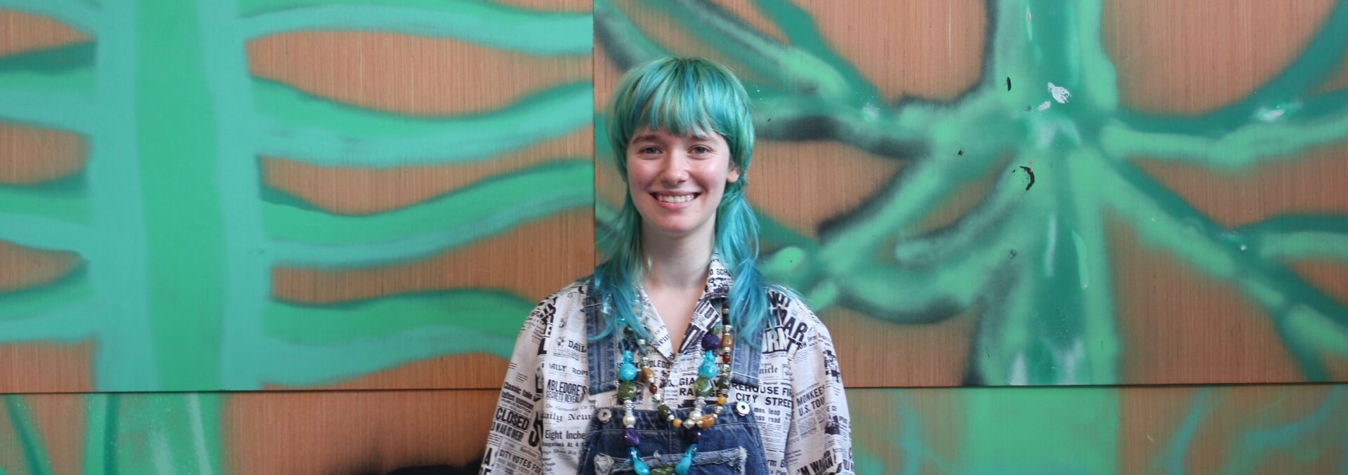 Photograph of volunteer Ella. She is wearing blue denim overalls and an off-white shirt. She is stood up, hands by her sides. She has green hair in a choppy style with a fringe. her green hair matches the green of the artwork in the background, which covers the entire wall. The artwork is wooden panels with green spray paint on, in shapes similar to palm leaves or green spines.
