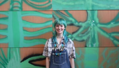 Photograph of volunteer Ella. She is wearing blue denim overalls and an off-white shirt. She is stood up, hands by her sides. She has green hair in a choppy style with a fringe. her green hair matches the green of the artwork in the background, which covers the entire wall. The artwork is wooden panels with green spray paint on, in shapes similar to palm leaves or green spines.