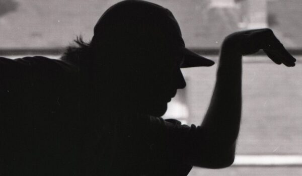 black and white image of Joe Hartley posing as a teapot as part of the Pos and Negs project. A silhouette, you can only see the outline of his body. Behind him is rooftops and chimneys.