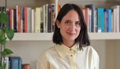 image of Kiera Blakey. She has shoulder length brunette hair, a cream coloured collared shirt. She is standing in-front of a book case populated with books of all colours. A monstera plant is in the far left of the image.