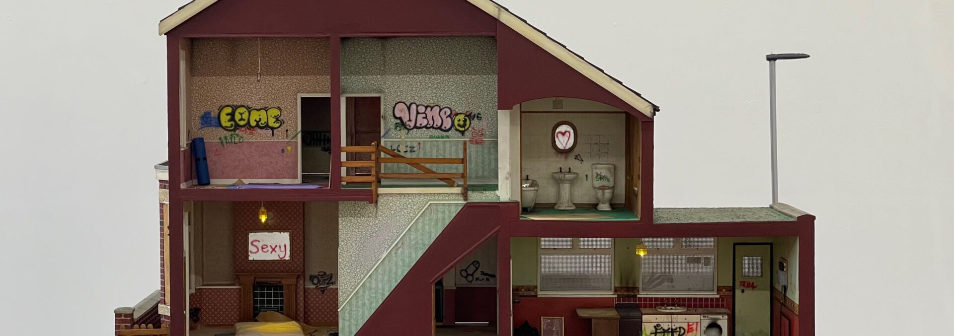 An artwork that is a toy model house, there are 3 rooms in the house and a basement garage. The artist has added detials within it