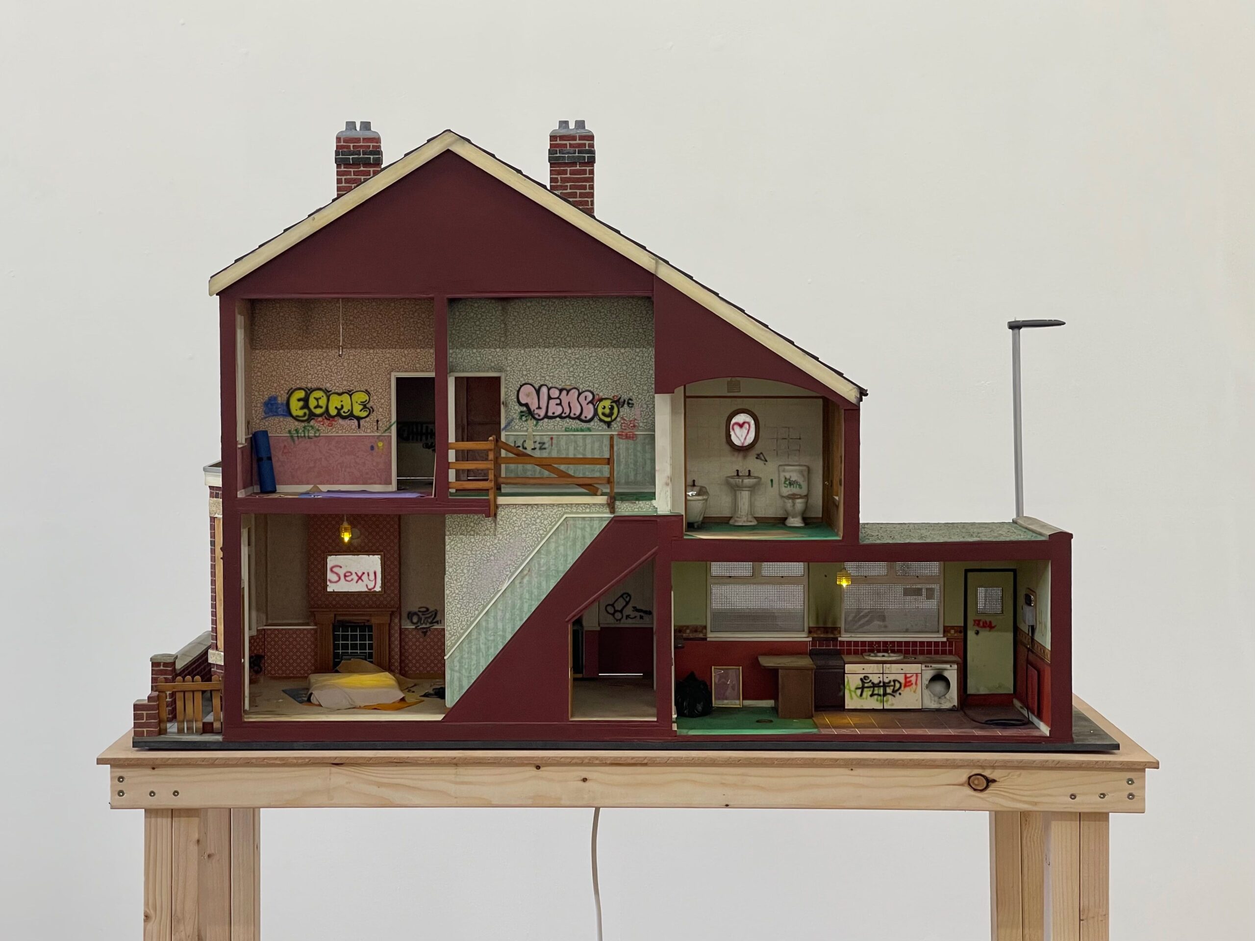 An artwork that is a toy model house, there are 3 rooms in the house and a basement garage. The artist has added detials within it