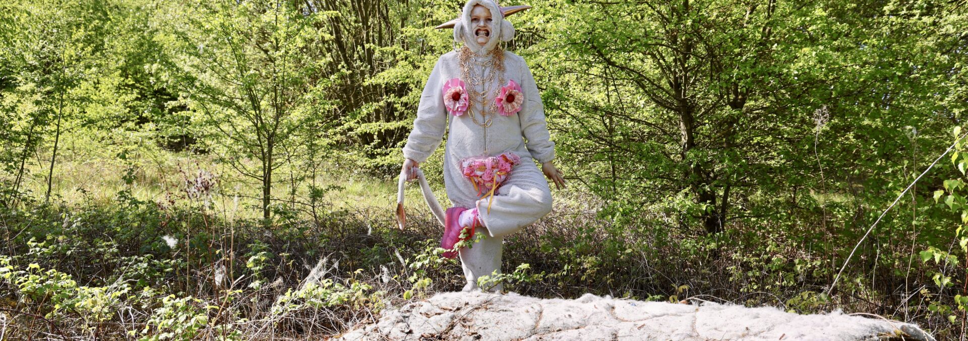 a woman dressed in a onesie with sewn on pink fabric, doing a dance in the middle of a field. There is a mattress and air spray bottle on the ground.