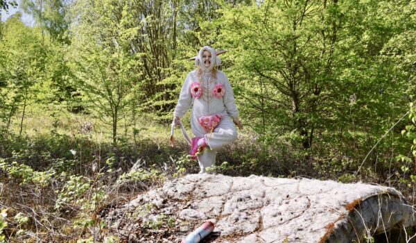 a woman dressed in a onesie with sewn on pink fabric, doing a dance in the middle of a field. There is a mattress and air spray bottle on the ground.