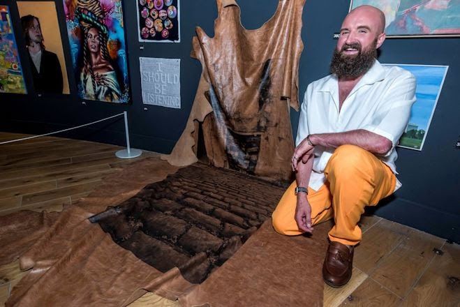 A Man Knealt next to his fabric artwork which hangs from the wall to the floor.
