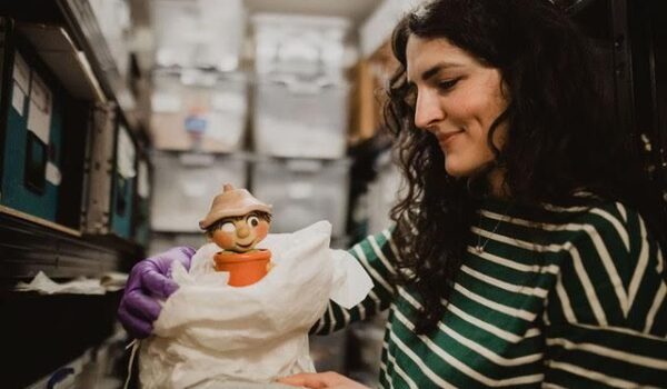 a woman with long, curly brunette hair. She is holding a clay model of a cartoon character in a plant pot.