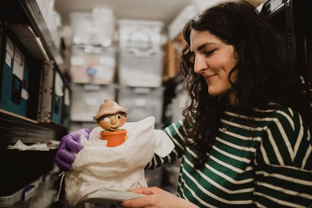 a woman with long, curly brunette hair. She is holding a clay model of a cartoon character in a plant pot.
