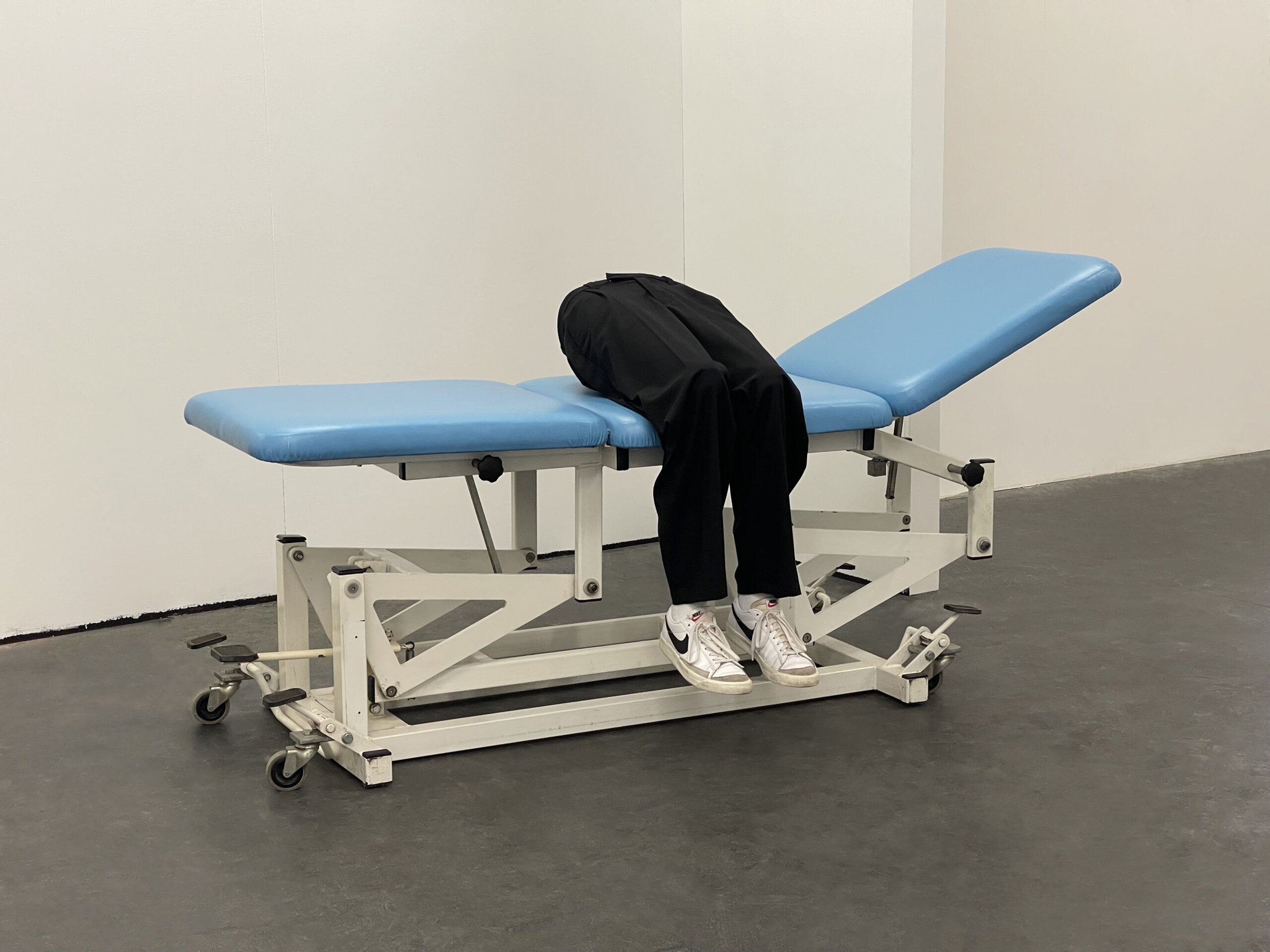 a photograph of a hospital bed with blue lining. A pair of legs wearing trousers and white trainers are sat on the bed. They appear to belong to a mannequin / dummy.