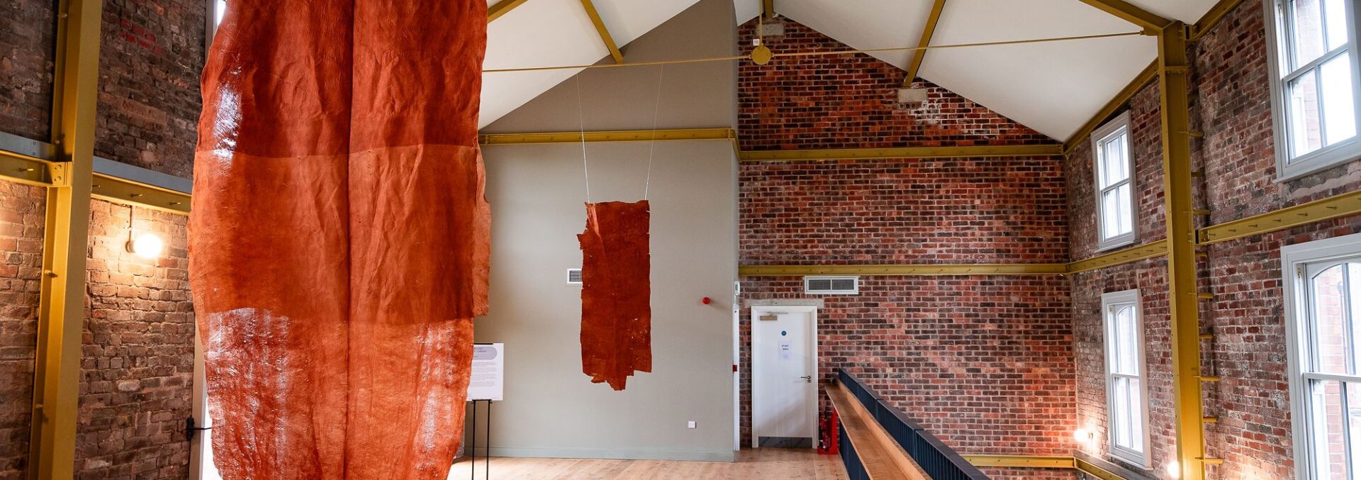 a large orange piece of fabtic hangs from a ceiling. It is in a large room, one side is white walls, the other side is exposed brick.