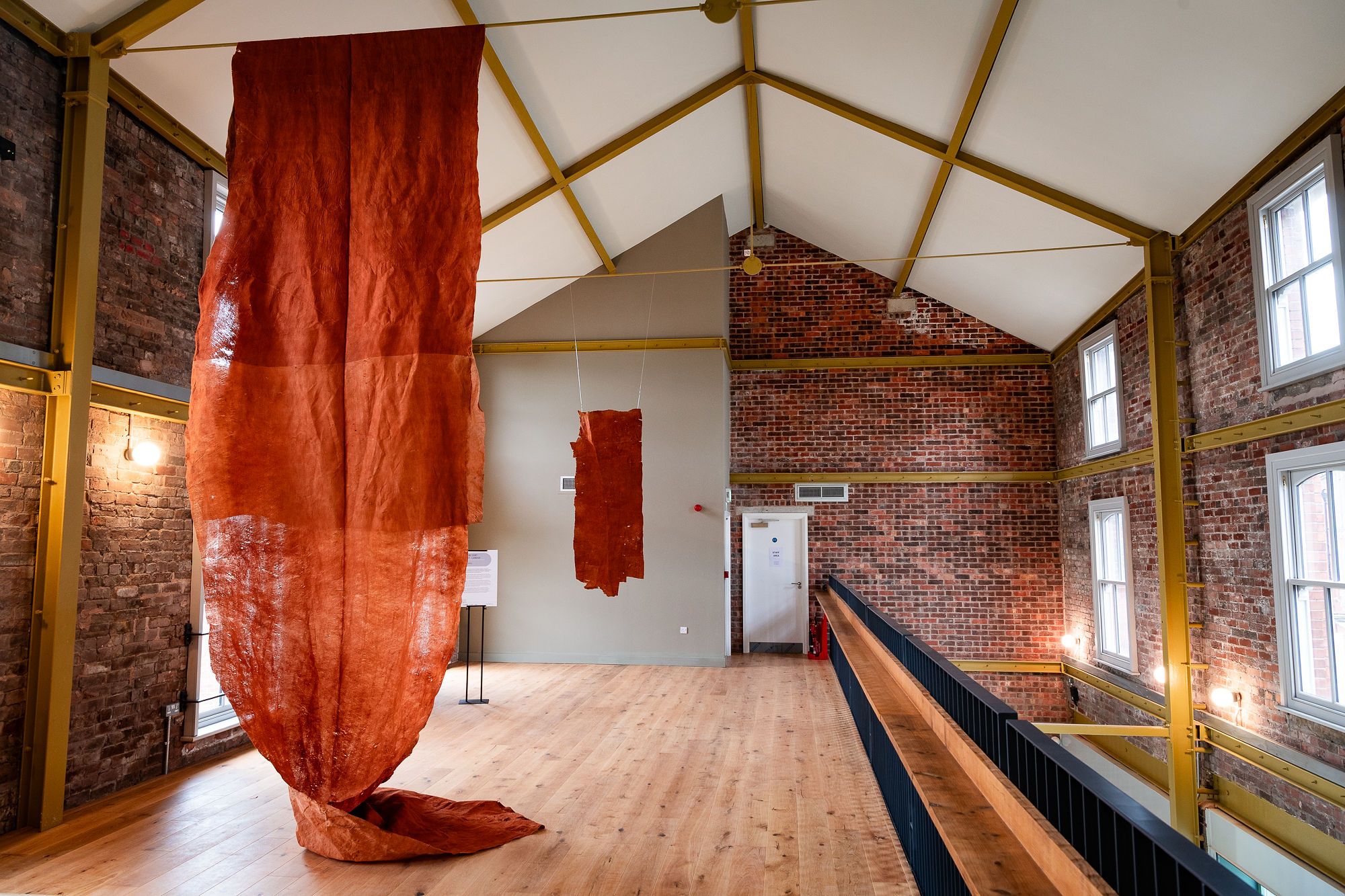 a large orange piece of fabtic hangs from a ceiling. It is in a large room, one side is white walls, the other side is exposed brick.
