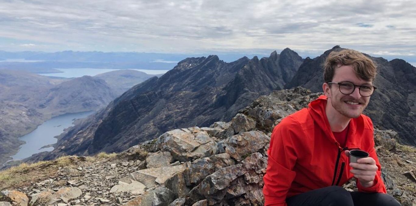 A young man smiling at the camera, he is wearing an orange jumper and crouching down. Behind him is a vas mountain range.