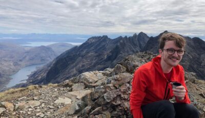 A young man smiling at the camera, he is wearing an orange jumper and crouching down. Behind him is a vas mountain range.