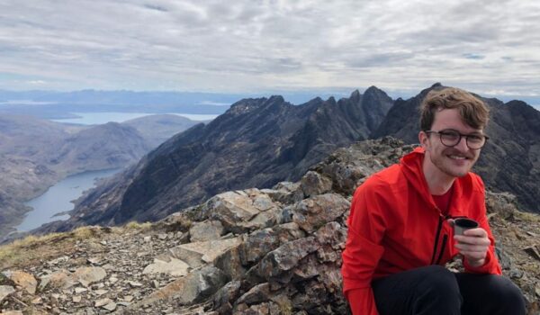 A young man smiling at the camera, he is wearing an orange jumper and crouching down. Behind him is a vas mountain range.