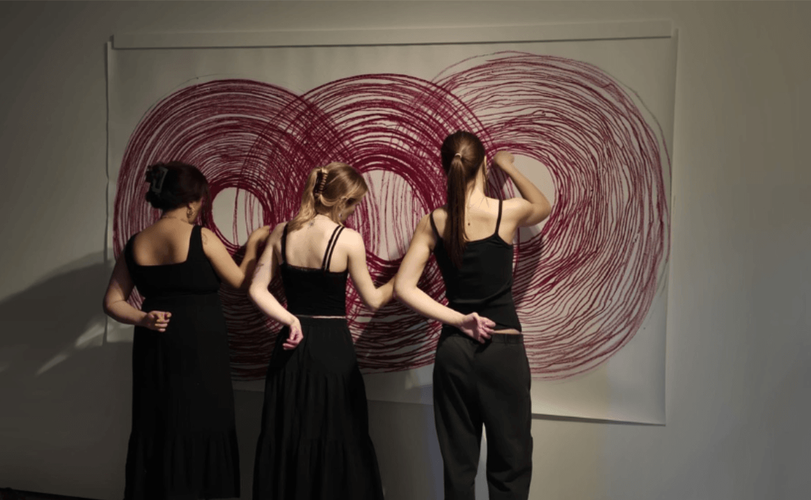 a photo of a live art performance, three women are drawing a sprial each, connected on a large piece of paper.