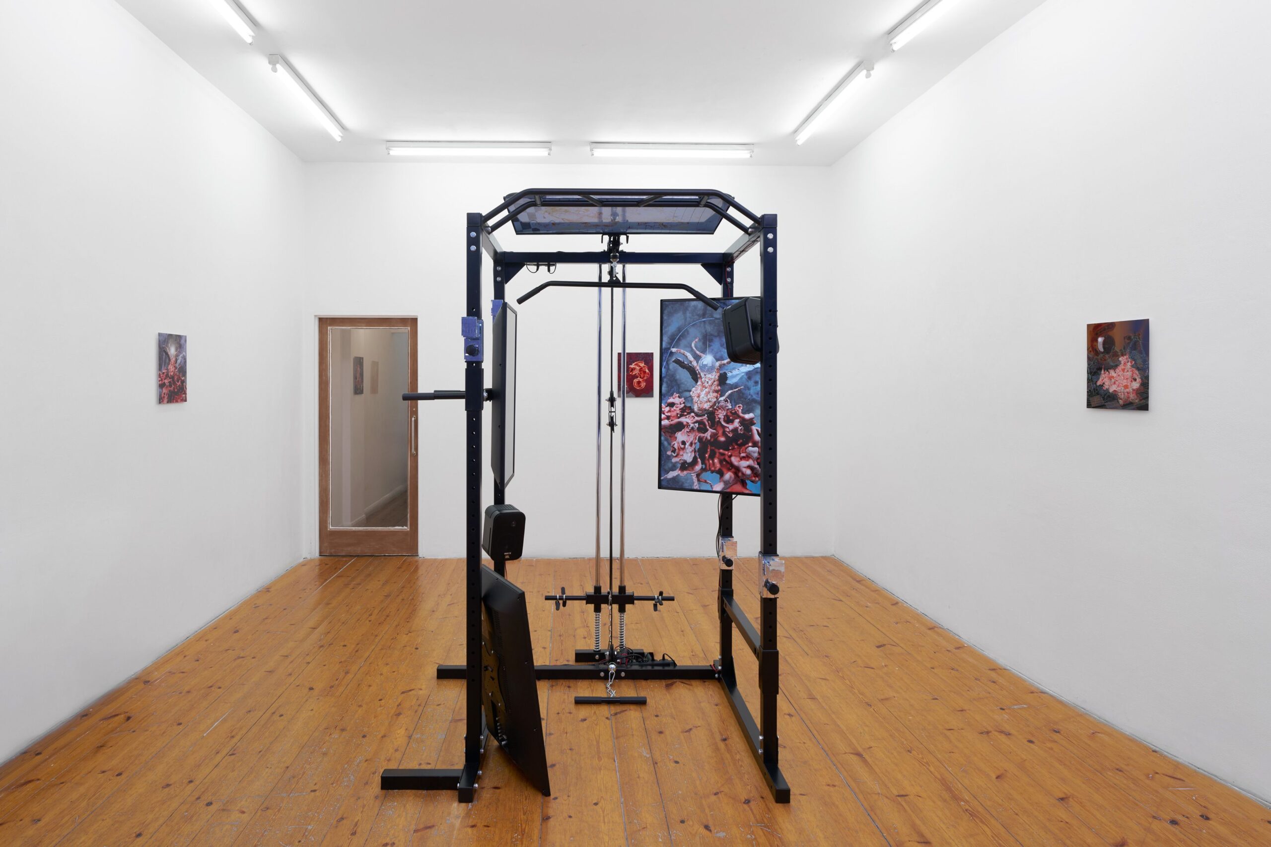 a small white walled gallery containing an art installation of a sculpture in the centre of the room made from black poles