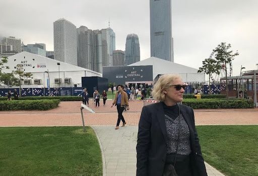 a woman with whispy blonde hair stood to the left of a photo with a city in the background.
